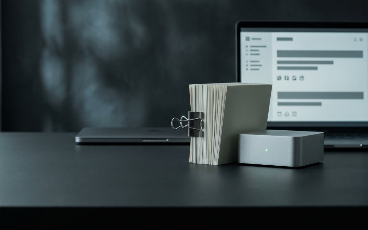 Стопка страниц под стальным зажимом, маленький серебристый сервер с зелёным огоньком и ноутбук